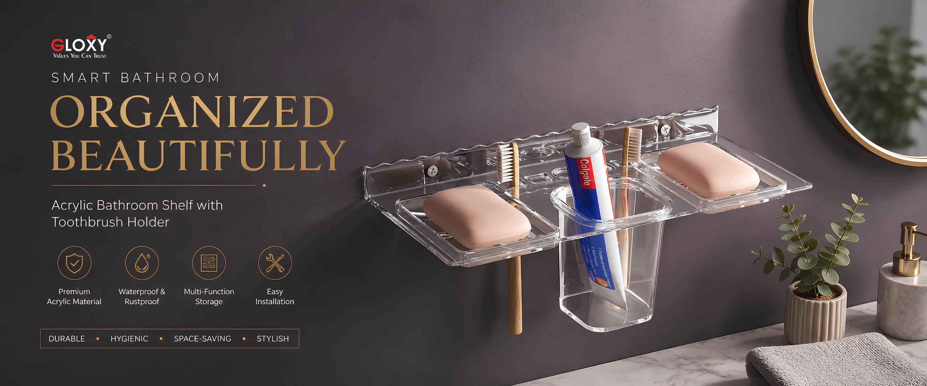 A wall-mounted acrylic bathroom organizer with two soap dishes and a toothbrush holder, shown installed on a dark bathroom wall with toothbrushes, soap bars, a mirror, and small plants. Text on the left reads 'GLXY SMART BATHROOM ORGANIZED BEAUTIFULLY' with feature icons below.