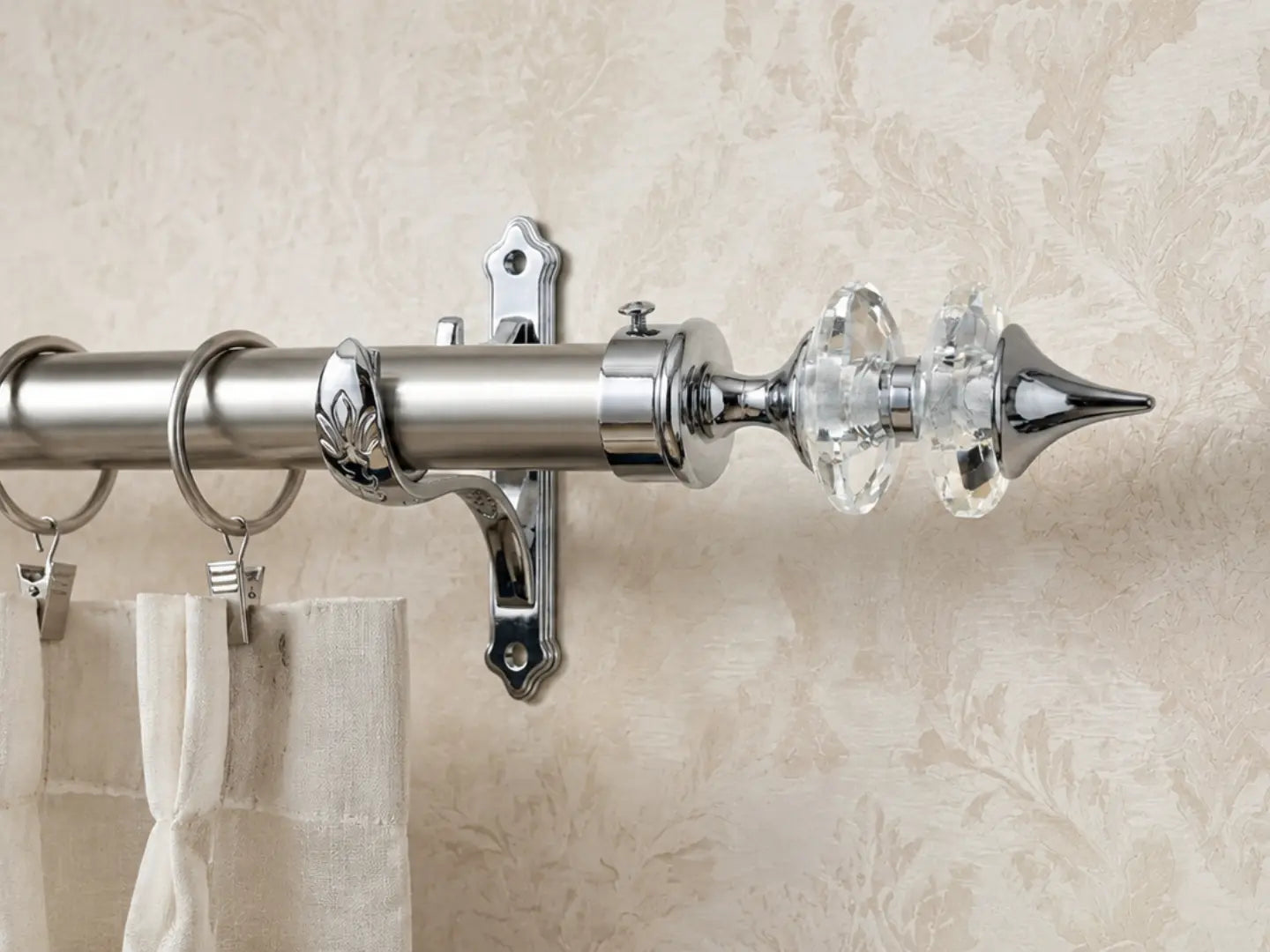A close-up of a polished silver curtain rod with ornate crystal finials and rings, mounted against a beige patterned wallpaper background.