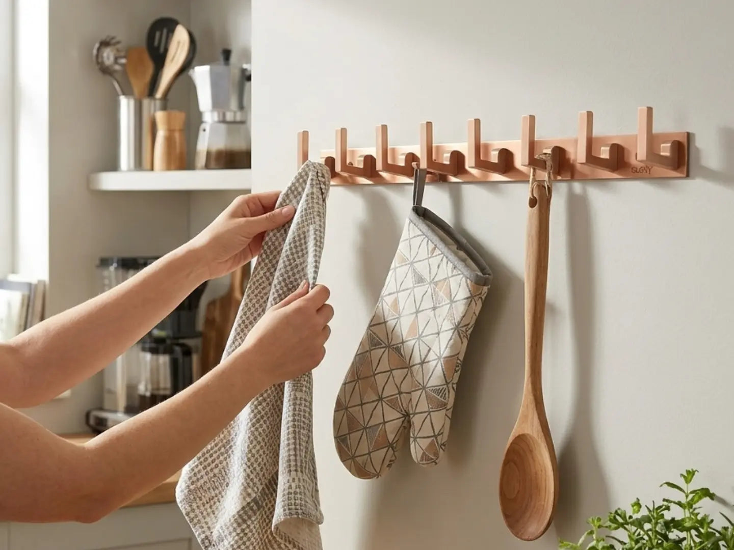 A wall-mounted wooden kitchen utensil rack with multiple hooks, shown in a bright kitchen setting. A person's hands are hanging a checkered kitchen towel, with a patterned oven mitt and a wooden spoon hanging on the rack.